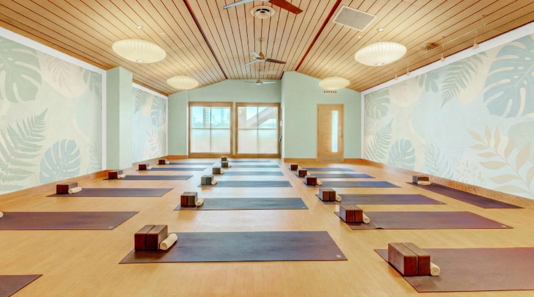A bright, empty yoga studio with polished wood floors and a vaulted wooden ceiling. Neat rows of dark yoga mats are laid out, each with two wooden blocks and a rolled white towel. The side walls are decorated with large, pale biophilic textures.
