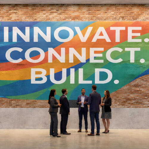 A large corporate mural on a brick wall featuring the words "INNOVATE. CONNECT. BUILD." in bold white text over a vibrant, colorful geometric wave pattern. Professionals in business attire are seen conversing and walking across a polished concrete floor.